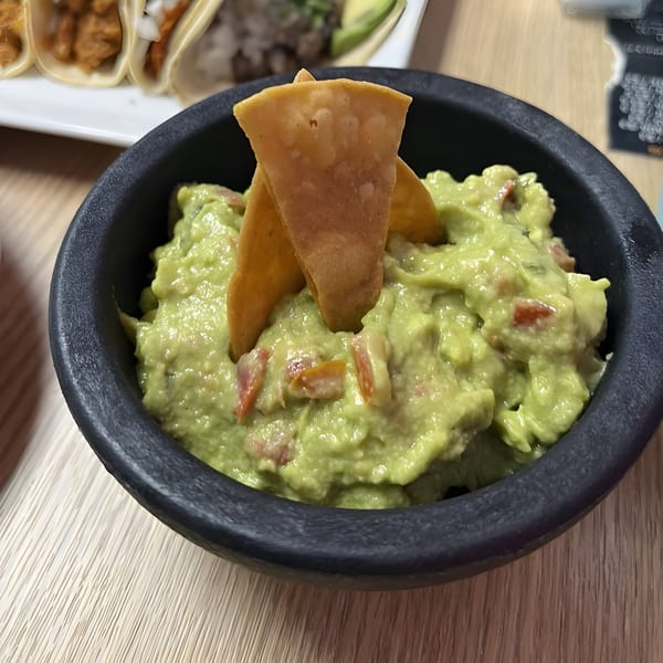 A black bowl of guacamole with two tortilla chips, on a wooden table with tacos in the background.
