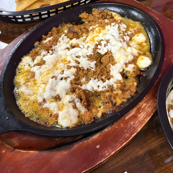 A sizzling skillet of melted white cheese and chorizo, with tortilla chips in the background.
