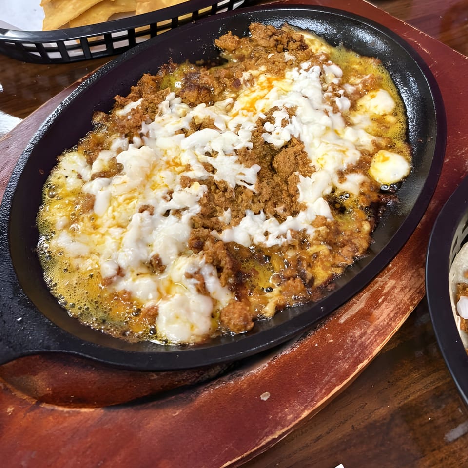 A sizzling skillet of melted white cheese and chorizo, with tortilla chips in the background.