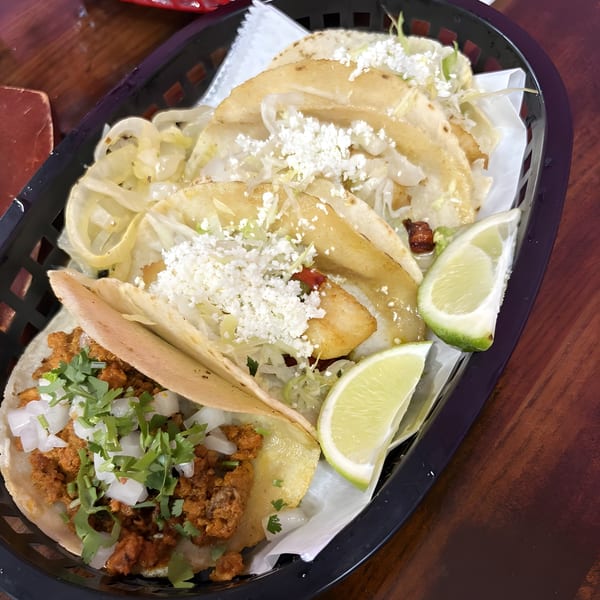 A black basket holds five Las Flores Mexican tacos with various fillings, cheese, and lime wedges.