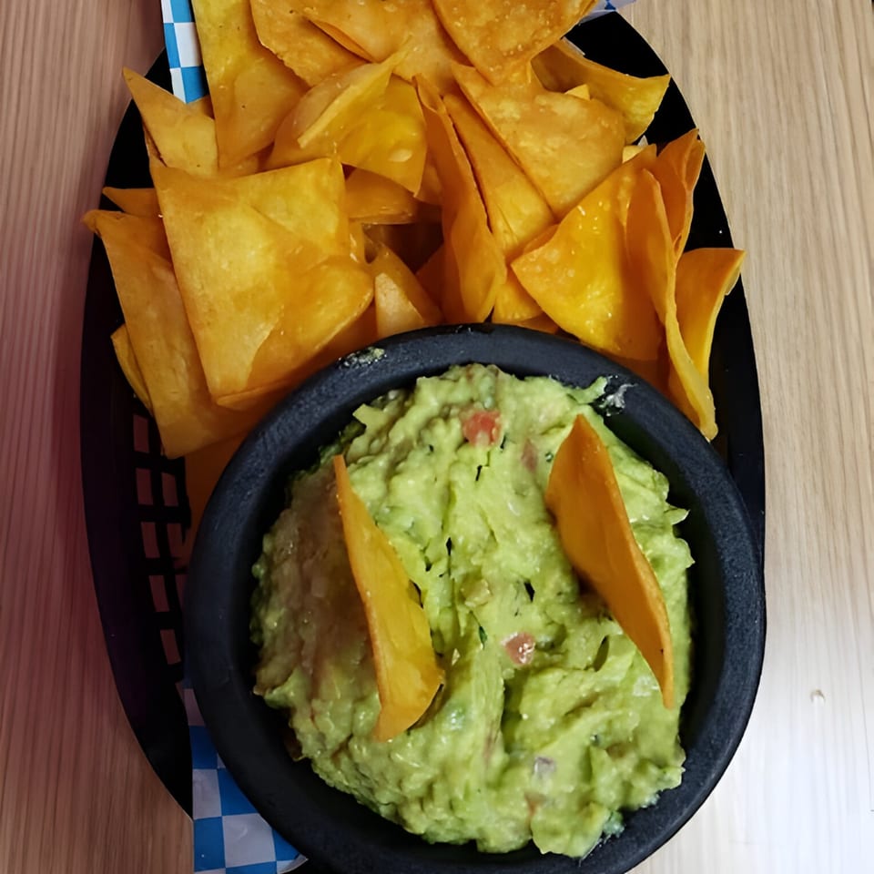 A black basket holds a bowl of guacamole with two chips, surrounded by a pile of tortilla chips.